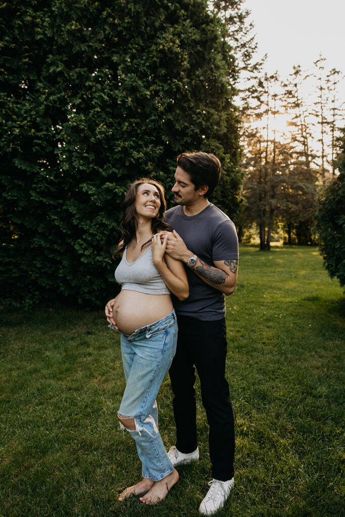Pregnant couple standing on the grass at sunset, her belly bare above unbuttoned jeans
