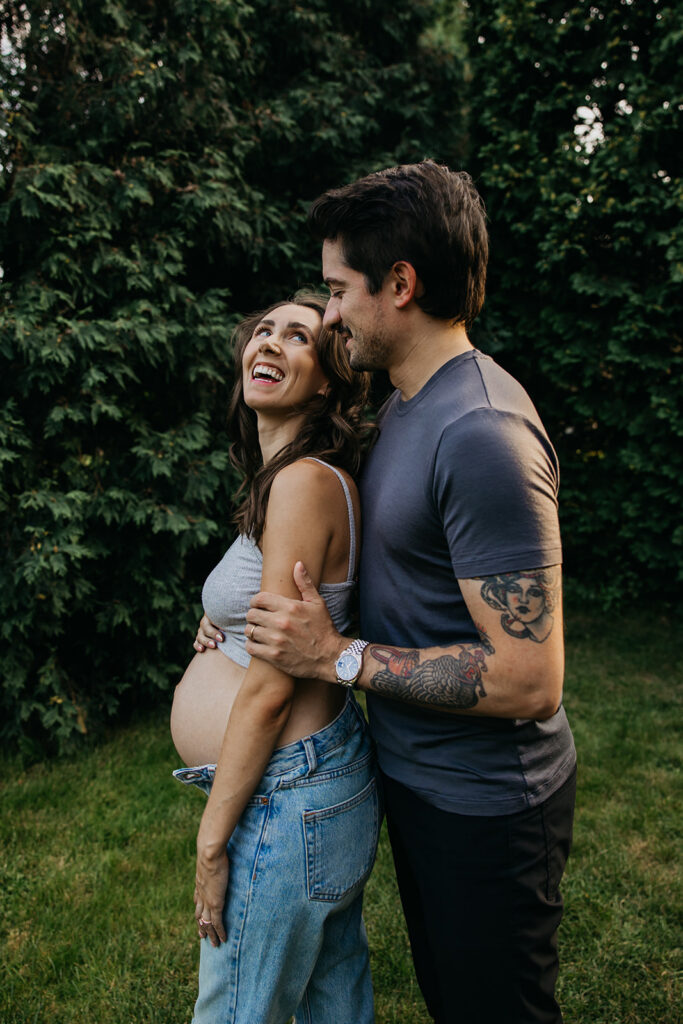 Pregnant woman laughing as her partner holds her close, tattooed arm around her belly