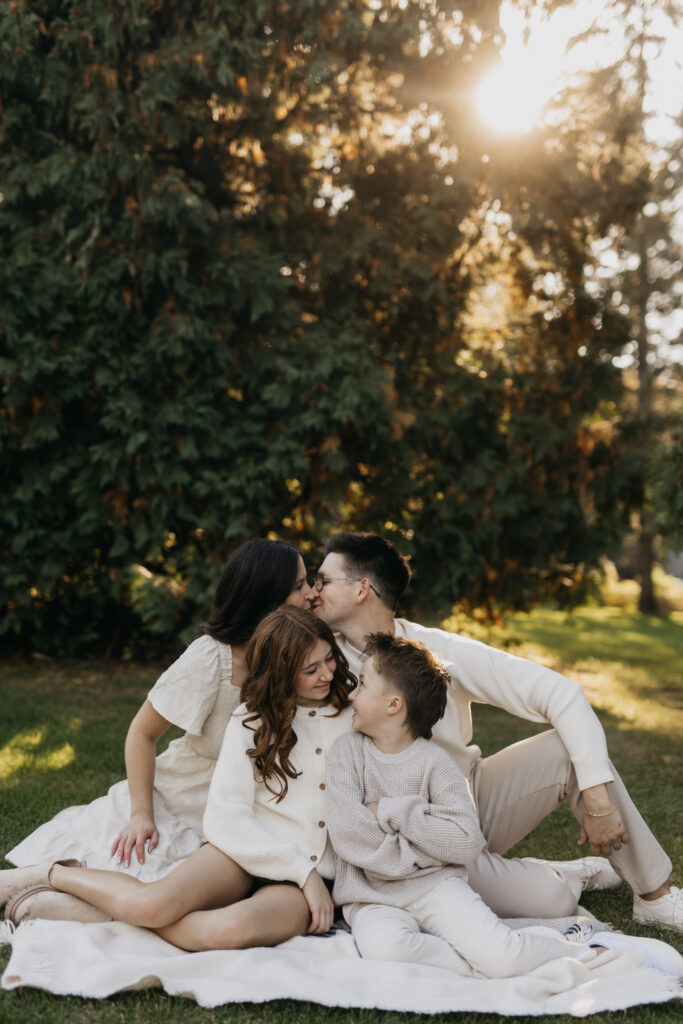 Mom and dad kissing while son and daughter lean in close together on a white blanket