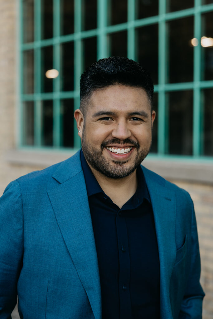 Man in a teal blazer and dark navy shirt smiling in front of green factory windows