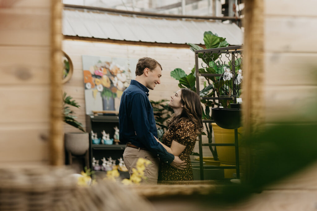 Couple holding each other inside a greenhouse surrounded by potted plants and yellow flowers