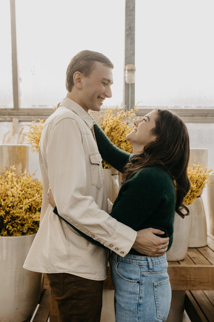 Couple embracing beside yellow flowers in a bright greenhouse during a winter engagement session