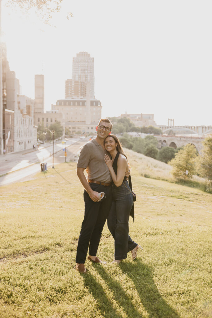 Newly engaged couple walking arm in arm across a Minneapolis hillside with the skyline behind them