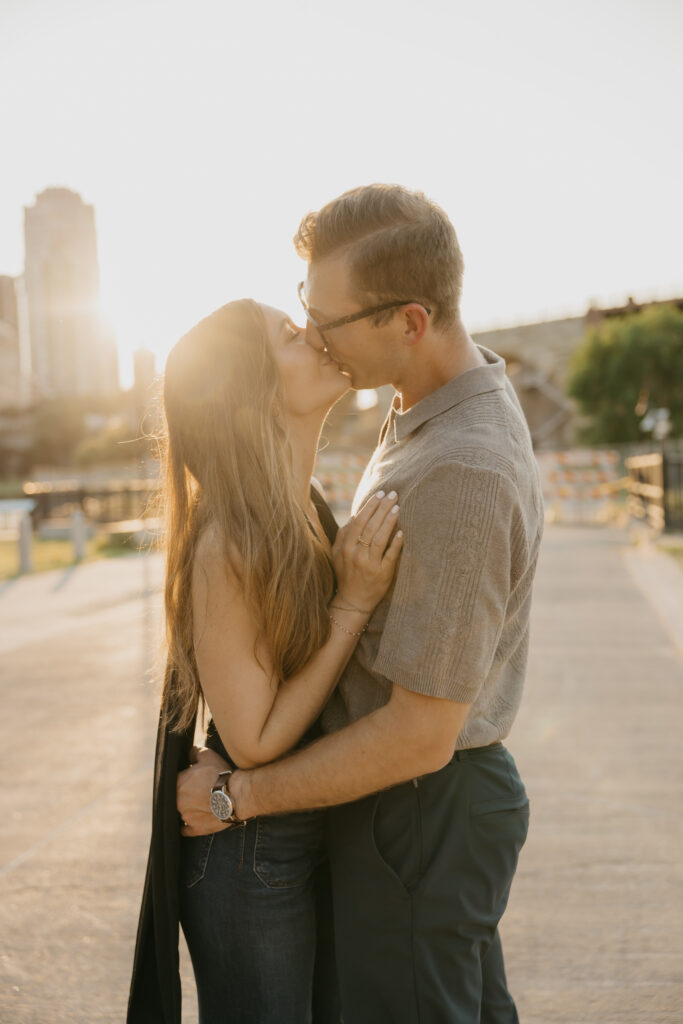 Engaged couple sharing a kiss backlit by the Minneapolis skyline at sunset
