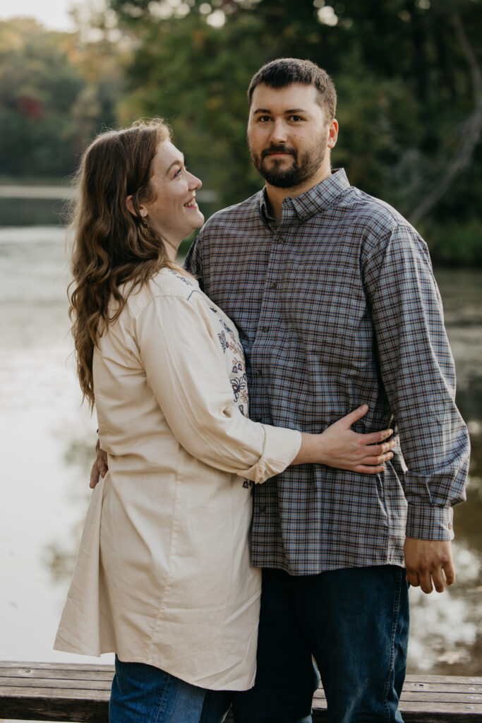 Couple standing arm in arm by the water with fall foliage reflected behind them