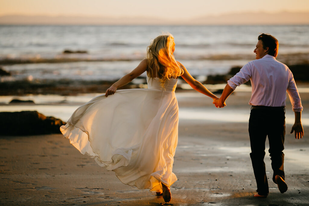 Couple running hand in hand along the shoreline at sunset with her dress flowing behind her