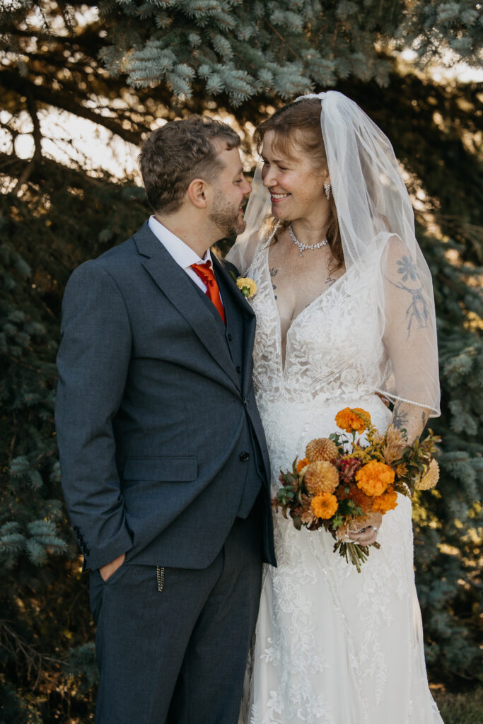 Bride and groom sharing a quiet moment beside pine trees with an orange bouquet