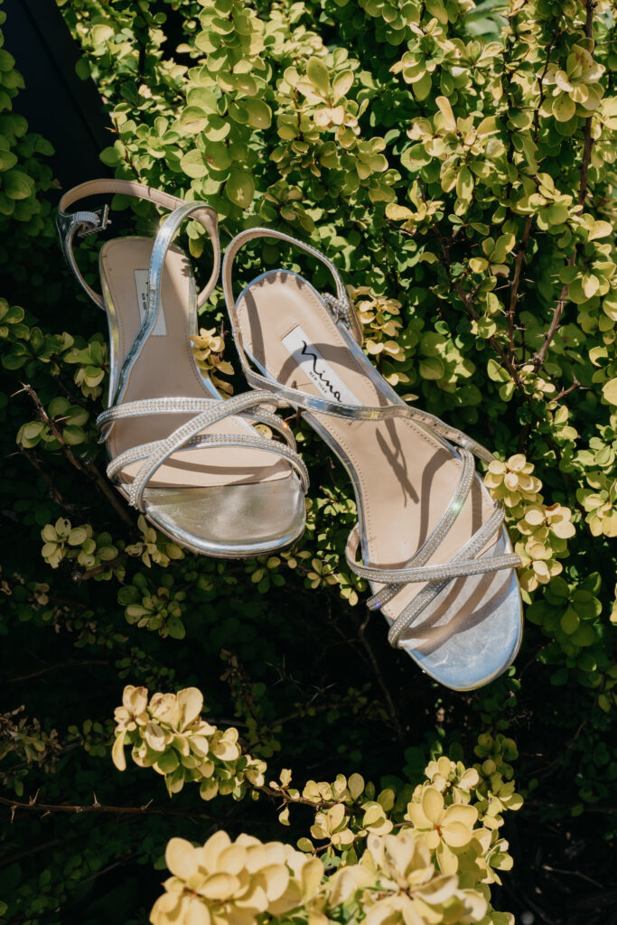 Silver strappy wedding heels hang in a hydrangea bush outside the ceremony venue