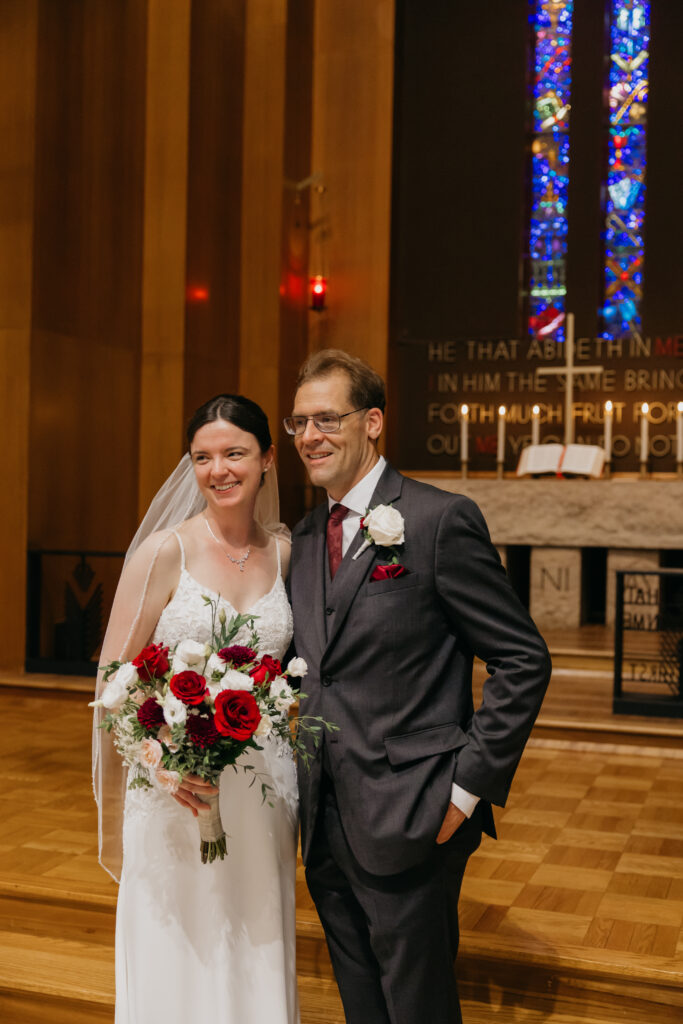 Bride and groom smile together at the altar holding their red and white bouquet