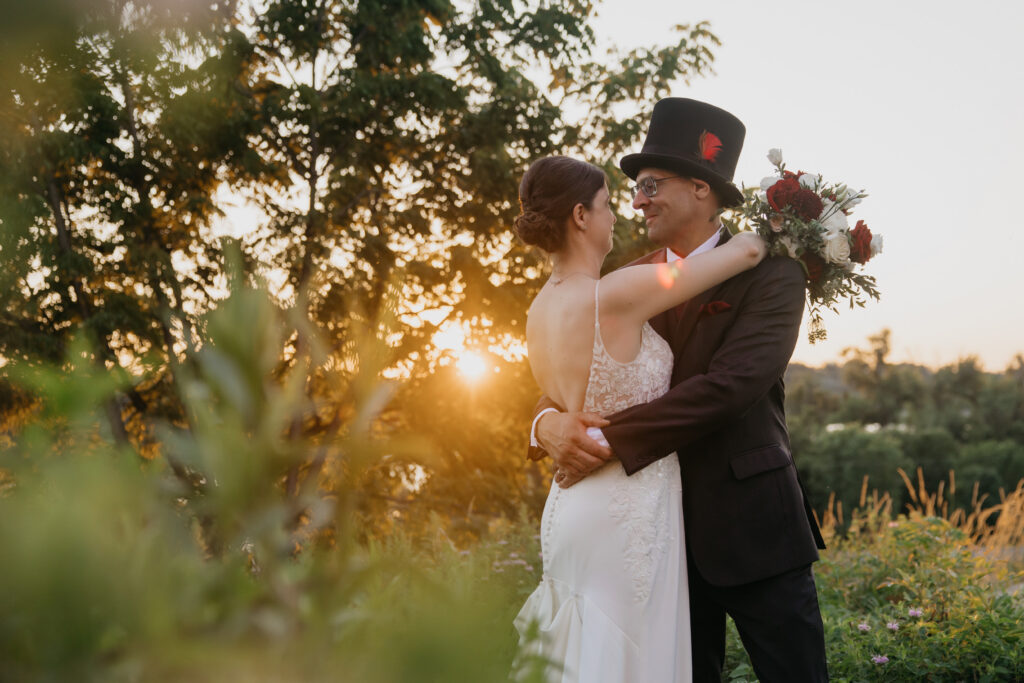 Bride and groom embrace closely in tall grass at sunset, groom in a black top hat