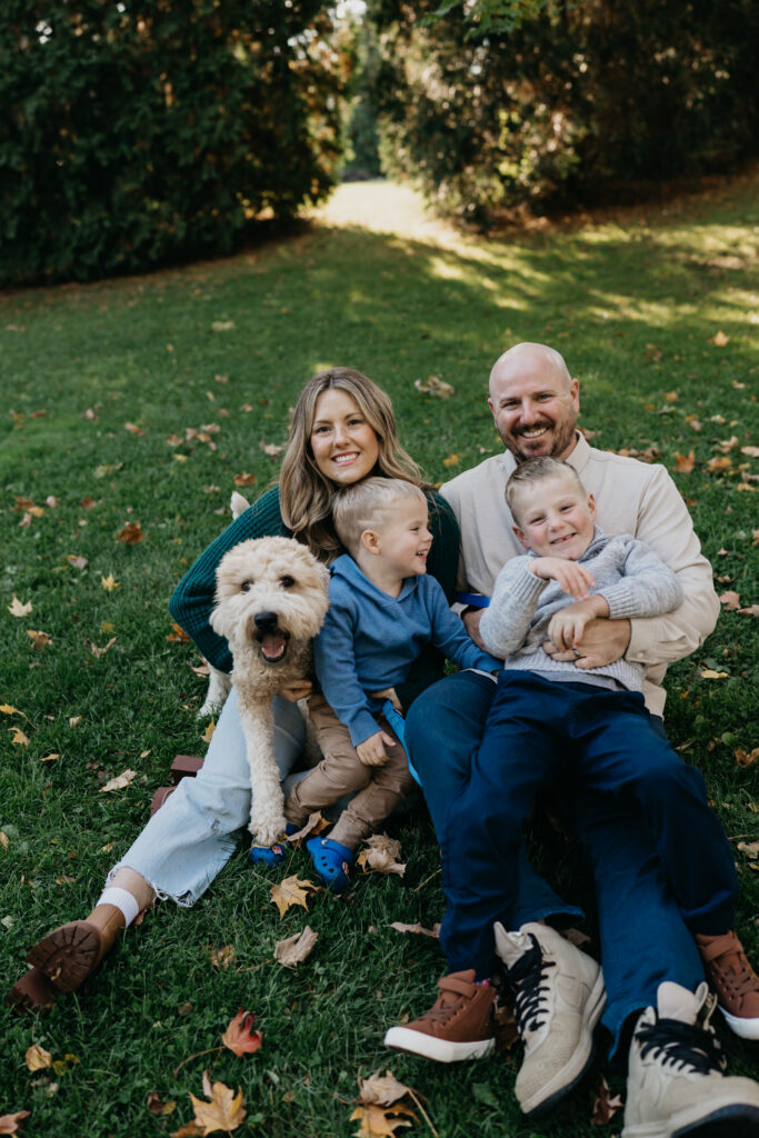 Family of four sitting on the grass with their goldendoodle among scattered fall leaves