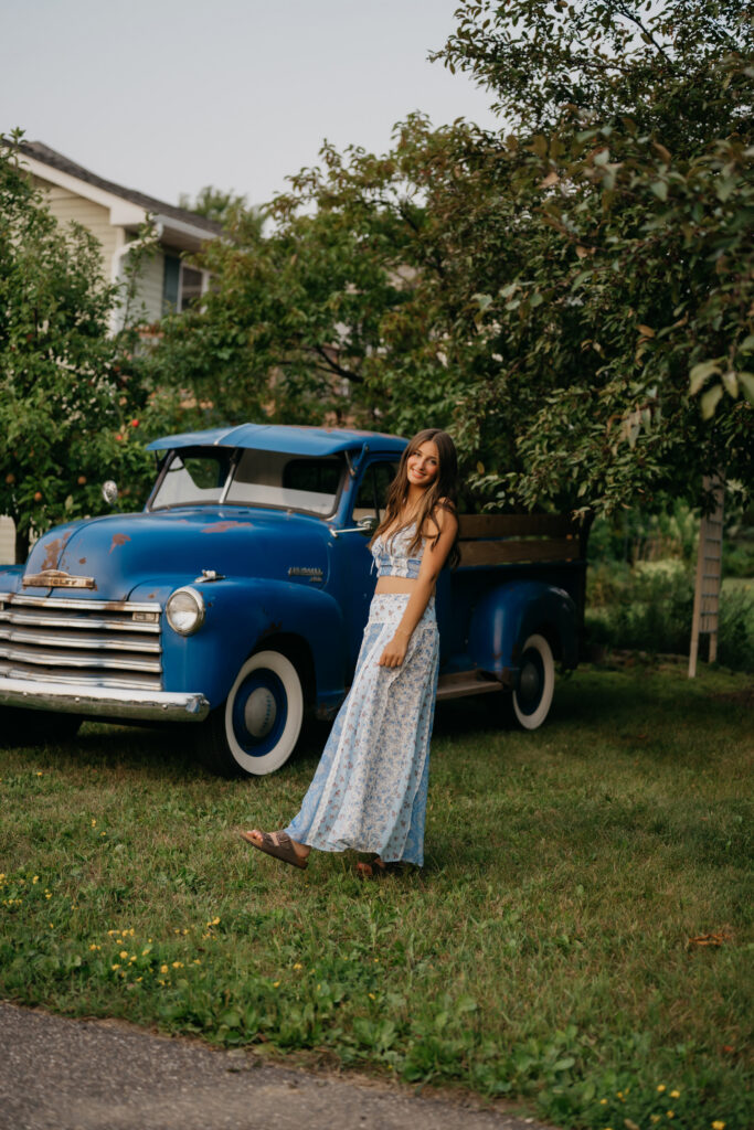 Senior in a blue floral maxi dress standing beside a vintage blue pickup truck