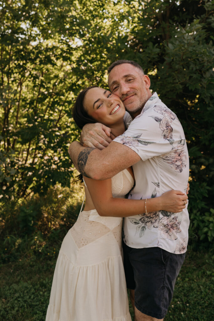 Senior girl in a white floral dress hugging her dad in a wooded backyard setting