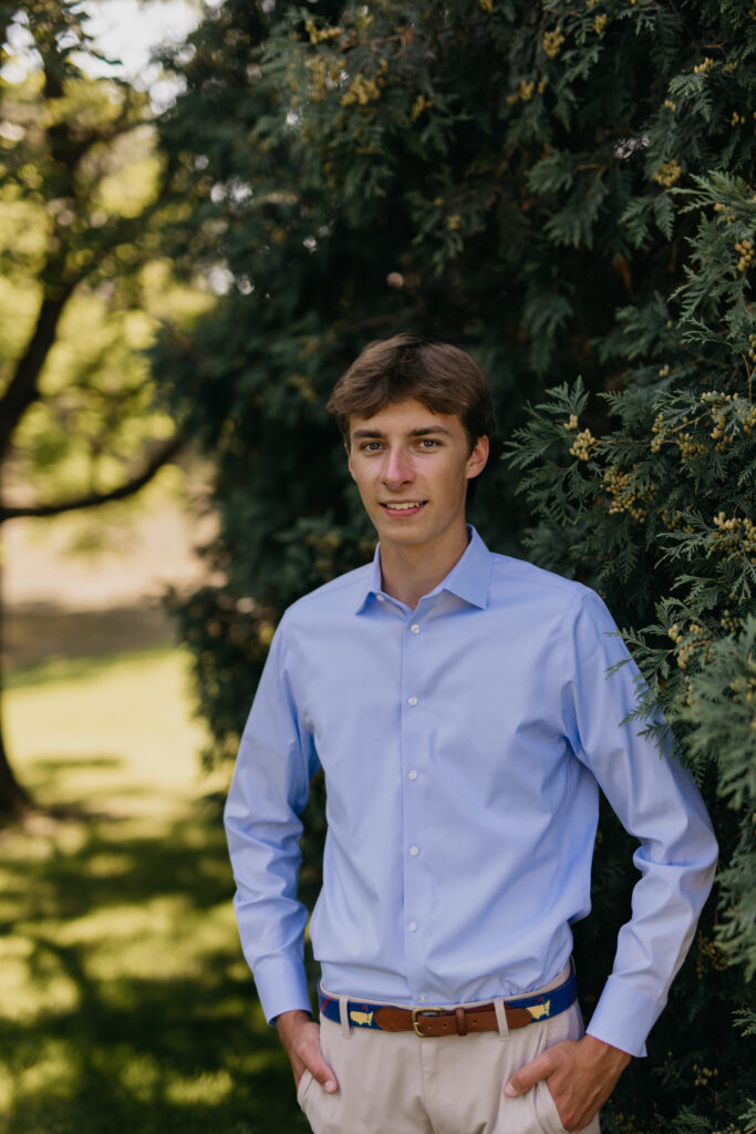 Senior guy smiling in a light blue button-up shirt in front of greenery