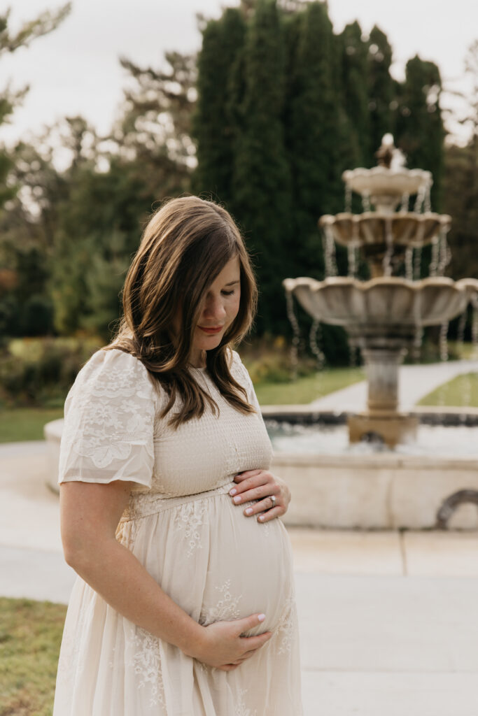 Pregnant woman in a cream dress looking down at her belly with a garden fountain behind her