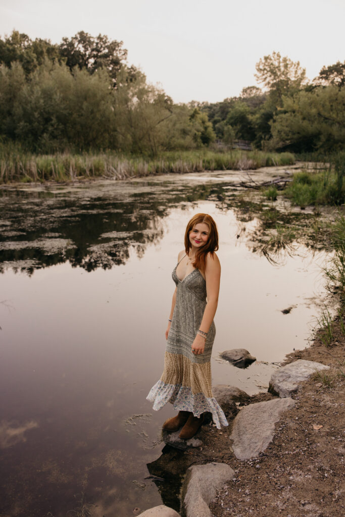 Senior in profile standing at the pond's edge with a soft water reflection behind her