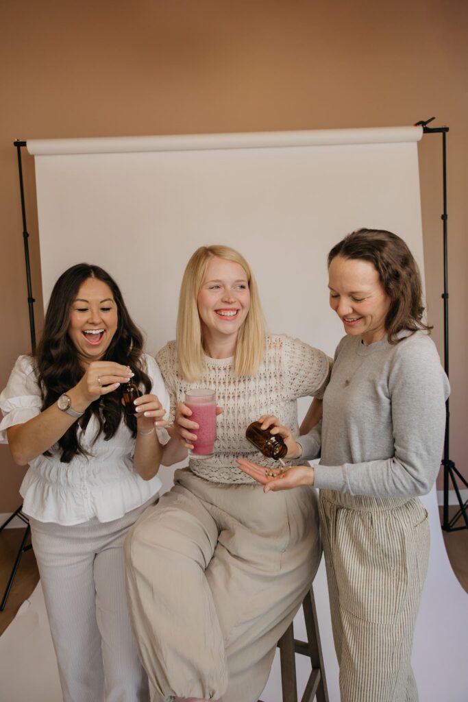 Three women holding a tincture dropper, pink smoothie, and supplements in front of a white backdrop