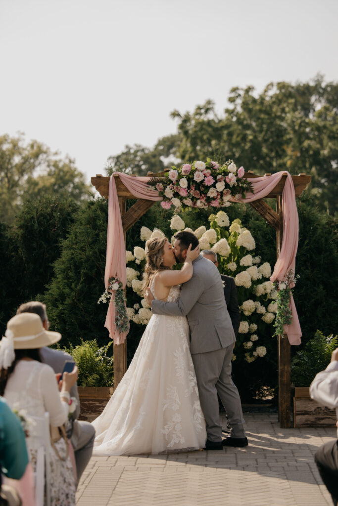 Bride and groom kiss at the altar under the floral arch as guests look on