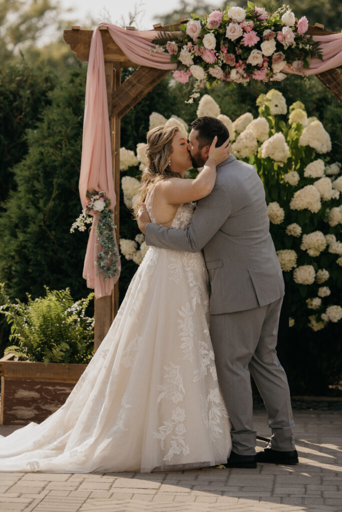 Bride and groom share their first kiss as husband and wife under a pink and white floral arch