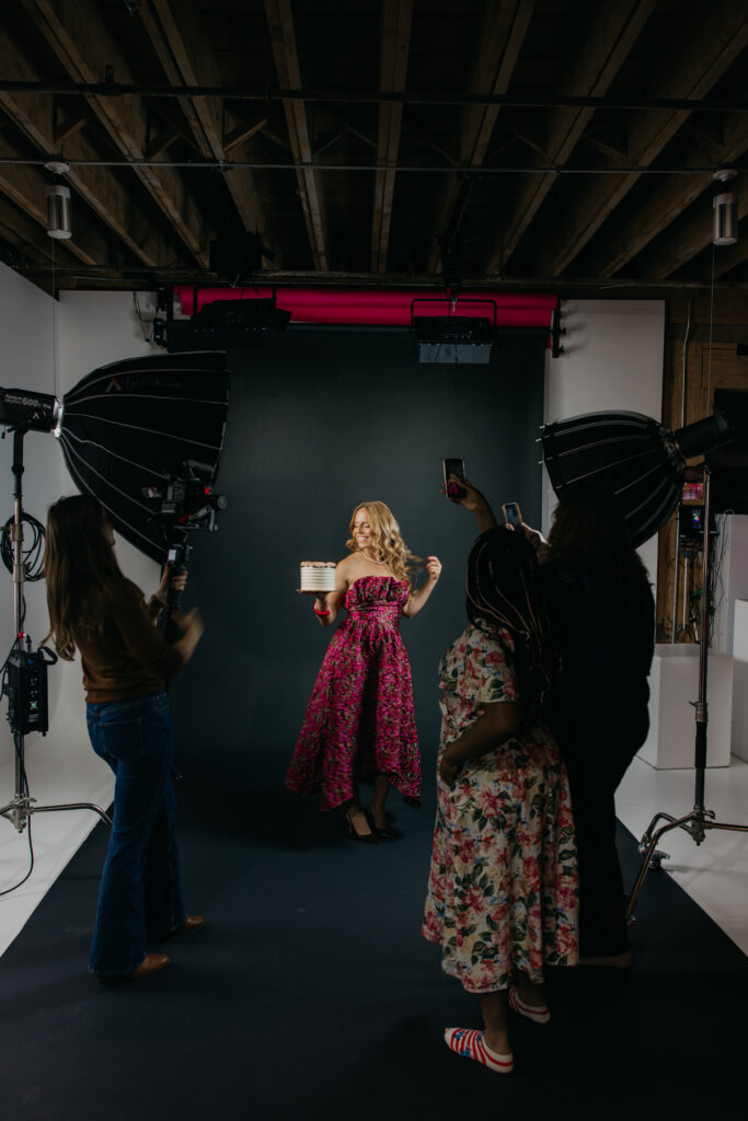 Creative business owner in a pink floral gown holding a small cake while her team photographs and films behind the scenes