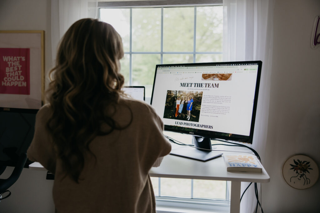 Creative business owner standing at her desk reviewing a "Meet the Team" webpage on her monitor