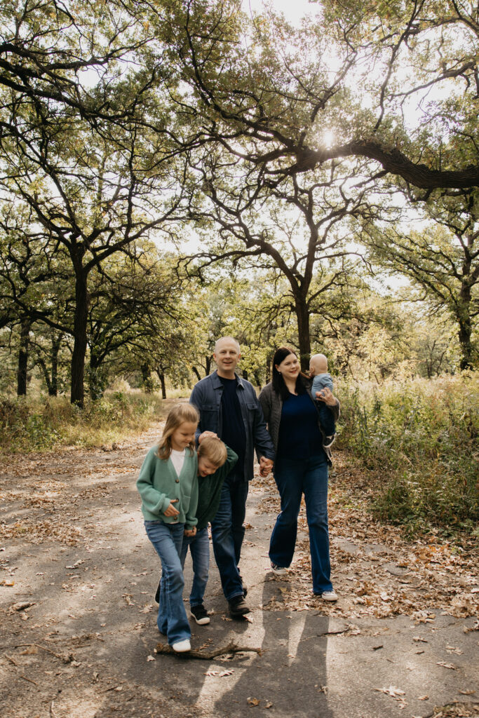 Family walking down a wooded trail, mom carrying baby as dad walks with two older children