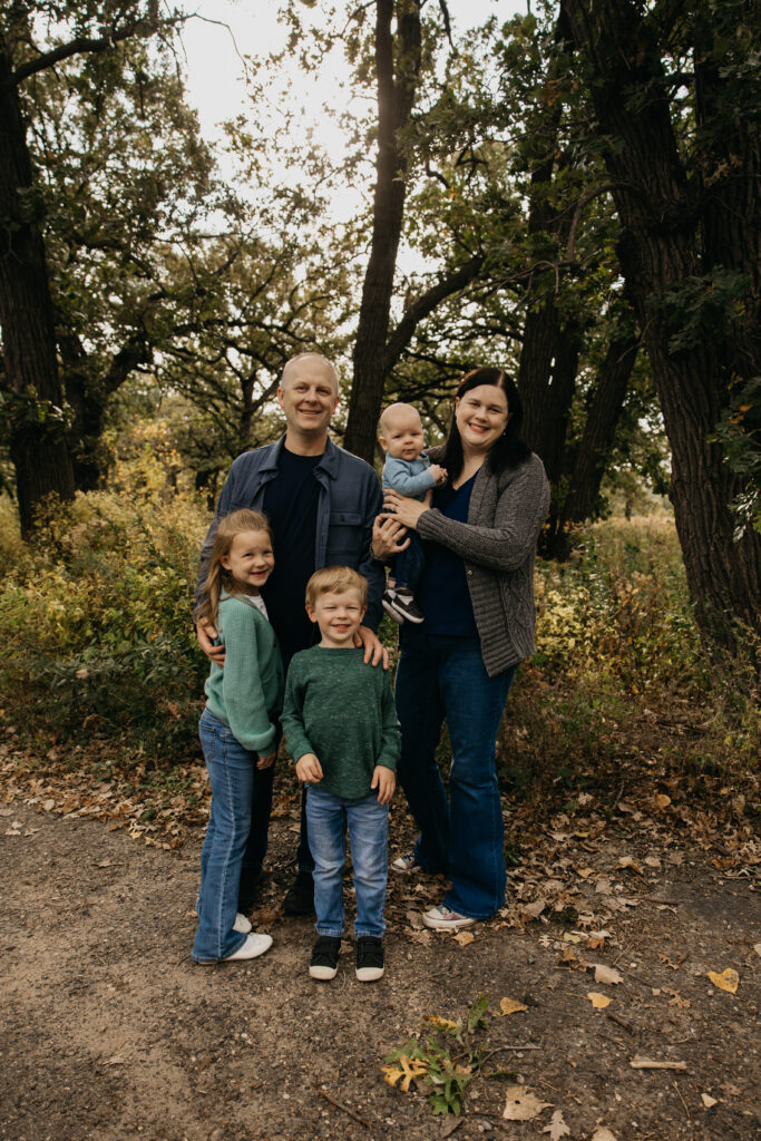 Family of five standing together on a dirt trail surrounded by tall oak trees
