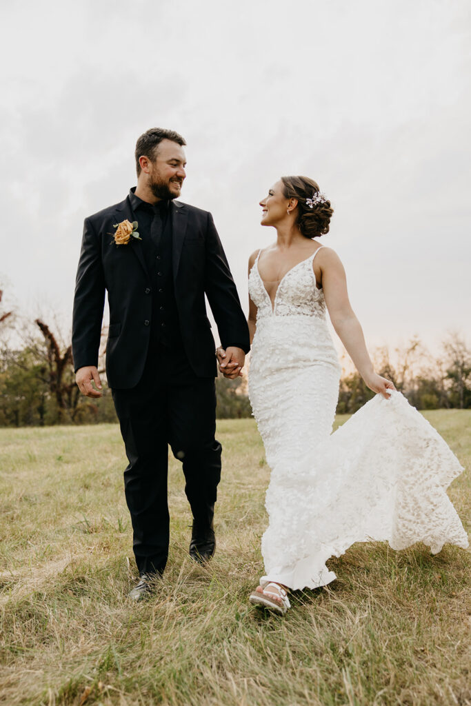 Bride and groom holding hands walking across a grassy hill at golden hour