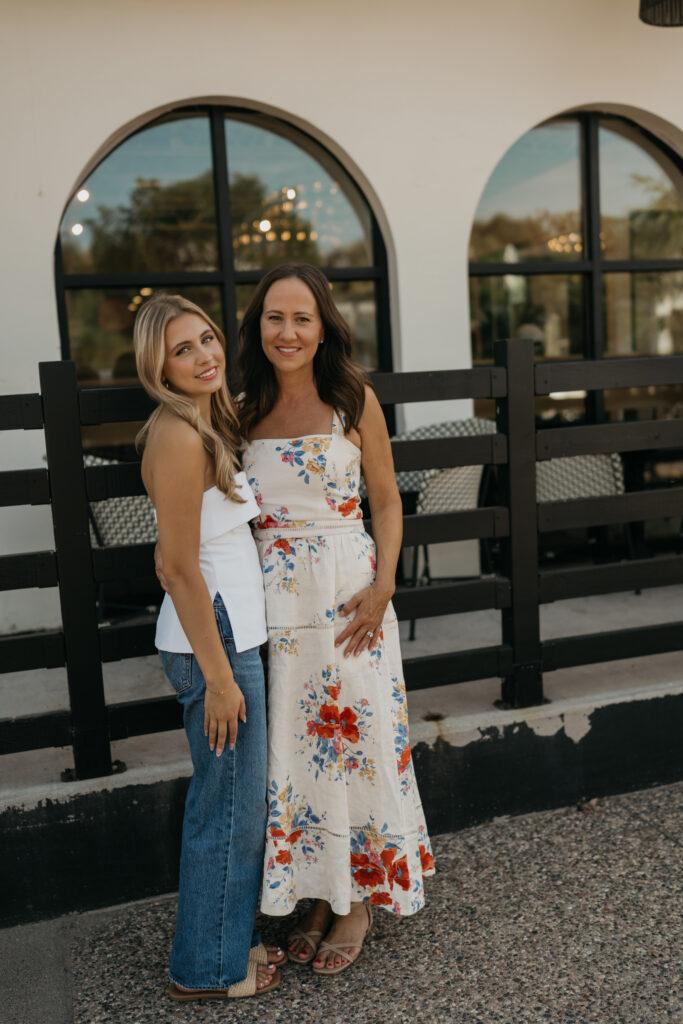 Senior girl in a denim skirt and mom in a floral maxi dress smiling together in front of arched windows