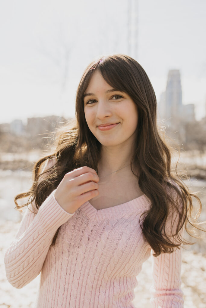 Senior girl in a pink sweater smiling with the Minneapolis skyline behind her