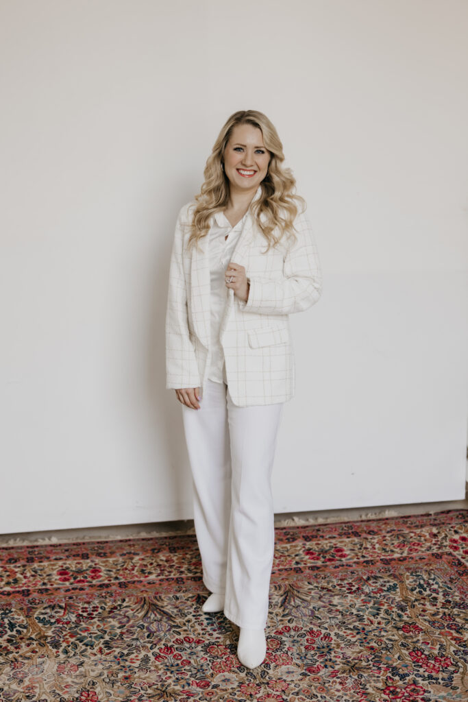 Blonde woman in a white textured blazer and white trousers smiling in a studio