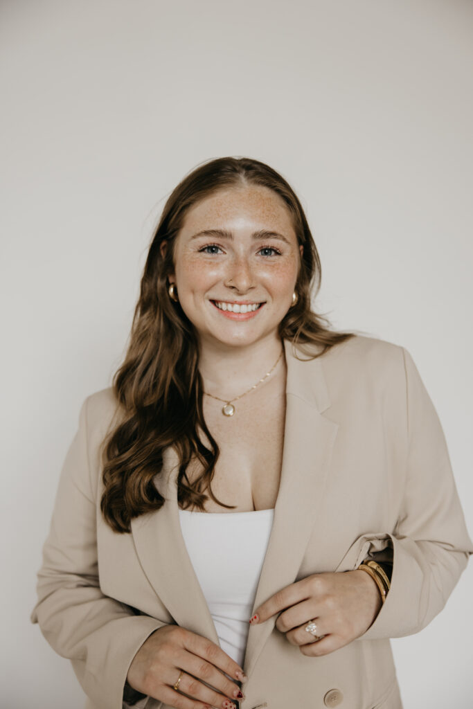 Woman in a beige blazer and white top smiling with gold jewelry in a studio headshot
