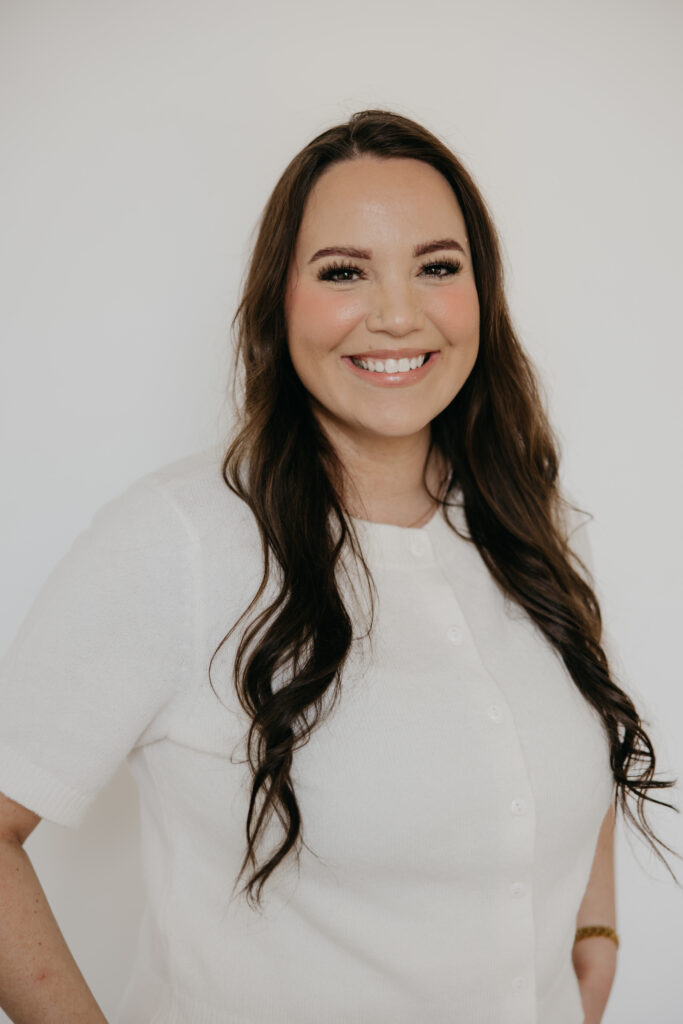 Woman with long dark hair in a white short-sleeve blouse smiling against a white backdrop