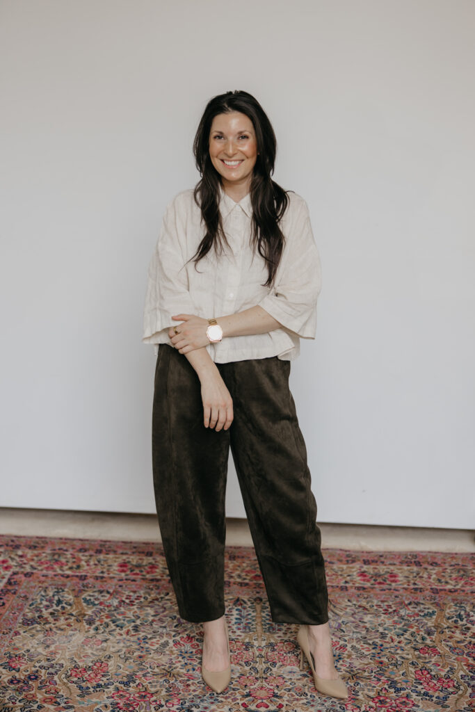 Woman in a cream button-up blouse and dark olive trousers smiling against a white studio wall