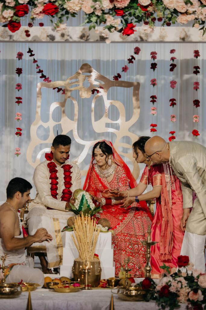 Priest leading a Hindu wedding ceremony under a white mandap with family gathered around the couple
