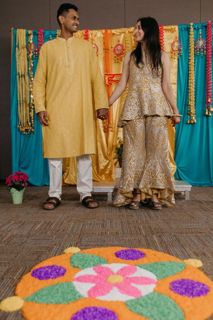 Couple in yellow and gold outfits holding hands before a colorful haldi backdrop and flower rangoli