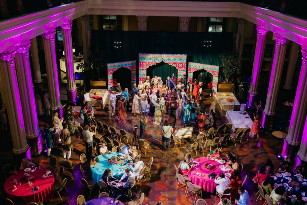 Overhead view of guests dancing in a grand pillared hall lit in pink during a sangeet