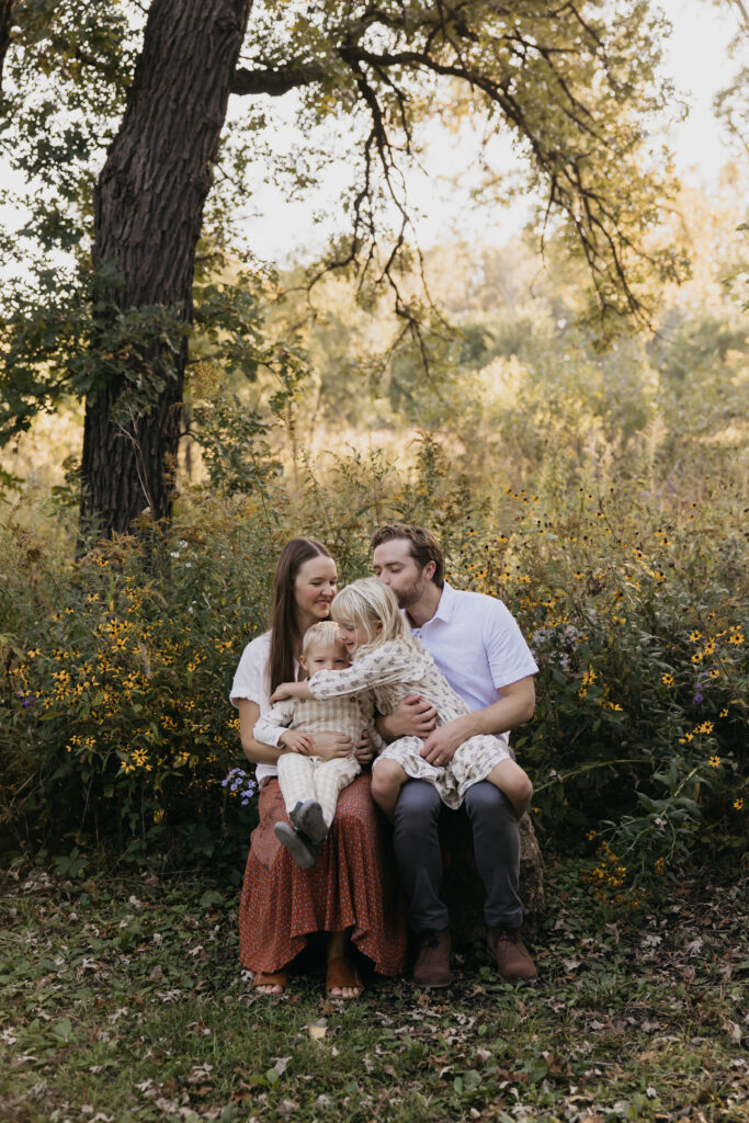 Mom and dad kissing toddler son's head while older daughter sits close by under a large tree