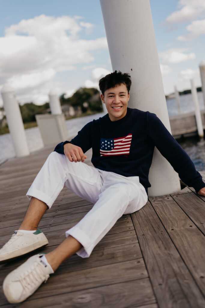 Smiling senior guy in a flag sweater and white pants lounging on a wooden dock