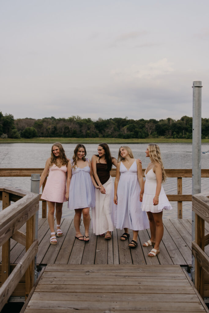 Group of five senior girls in pastel dresses walking together along a lakeside dock