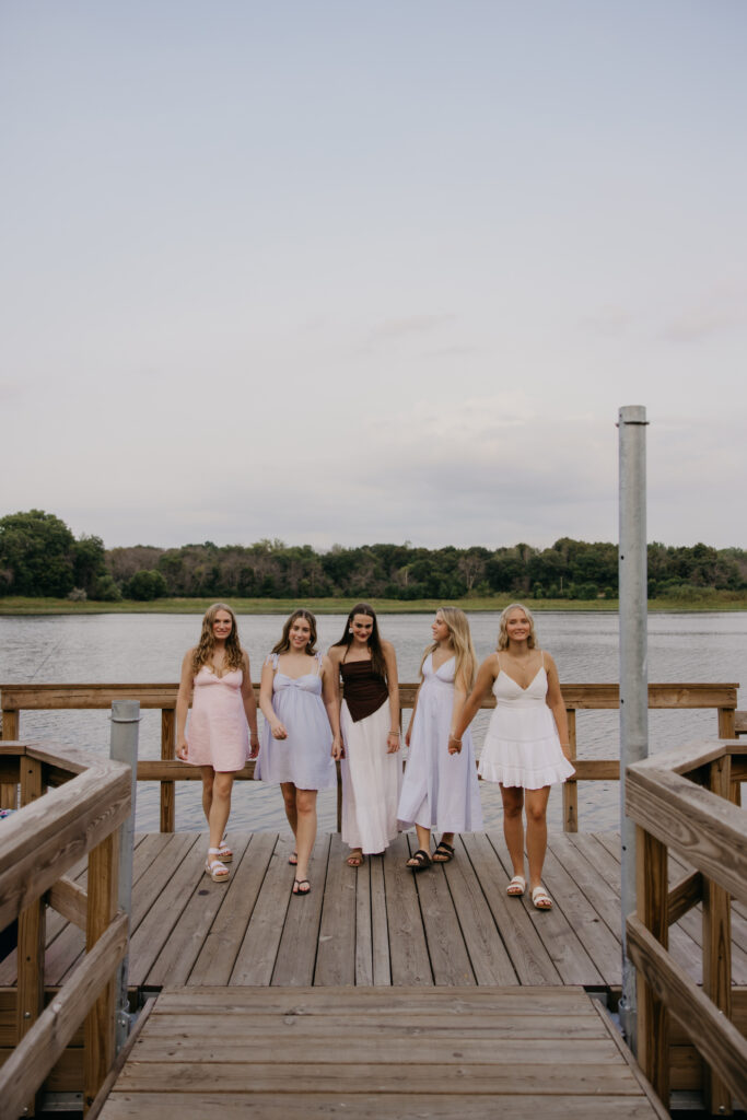 Group of five senior girls in pastel dresses walking hand in hand along a lakeside dock