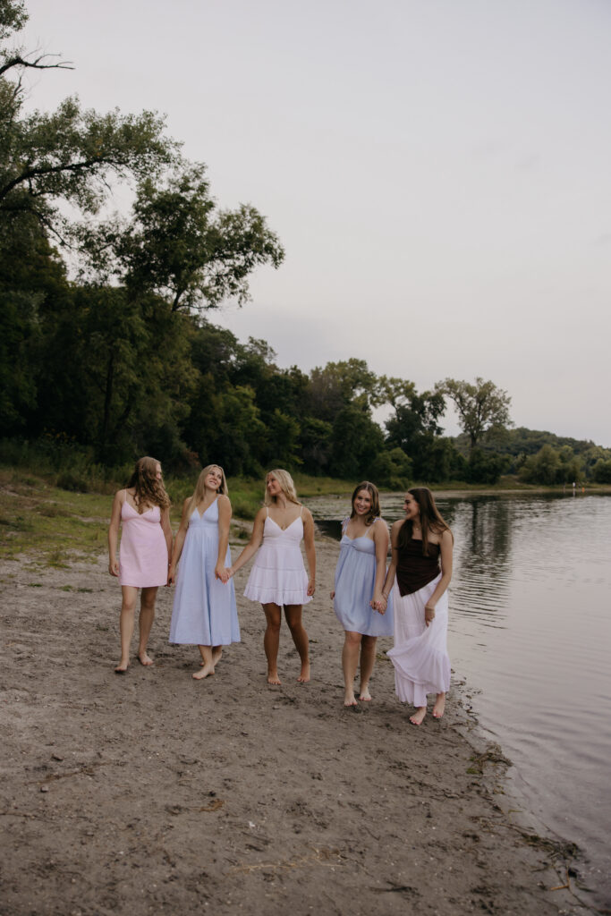Five senior reps in pastel dresses walking along a sandy lakeshore at sunset