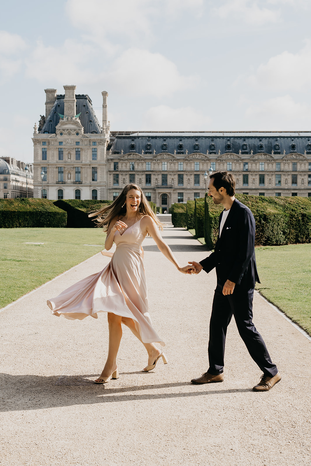 Engaged couple laughing and holding hands on a garden path in front of the Louvre in Paris