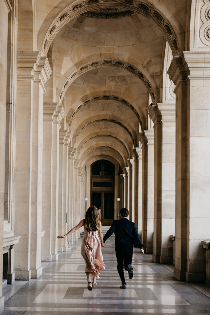 Engaged couple running hand in hand through a Paris colonnade
