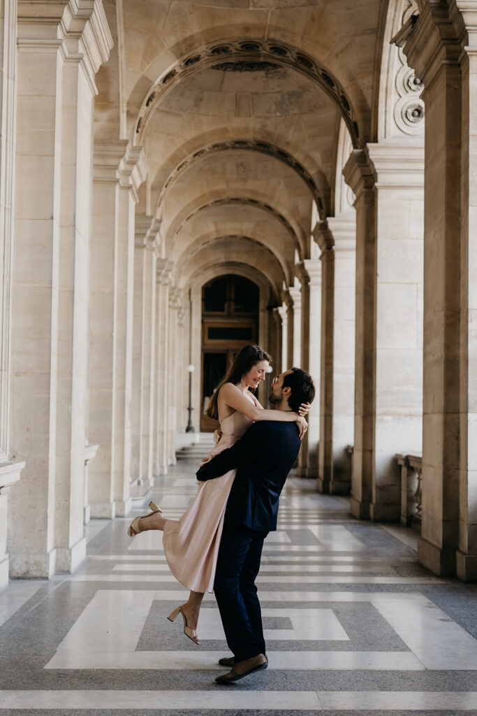 Man carrying his fiancée while she laughs, under a colonnade in Paris
