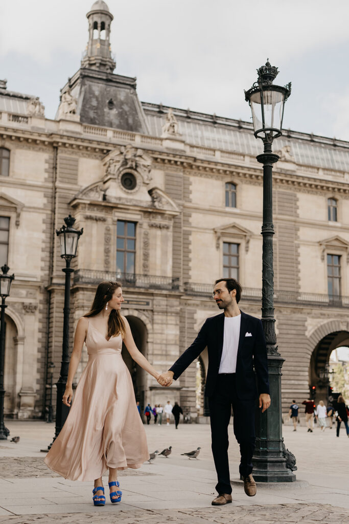Engaged couple holding hands and walking past pigeons in the Louvre courtyard in Paris