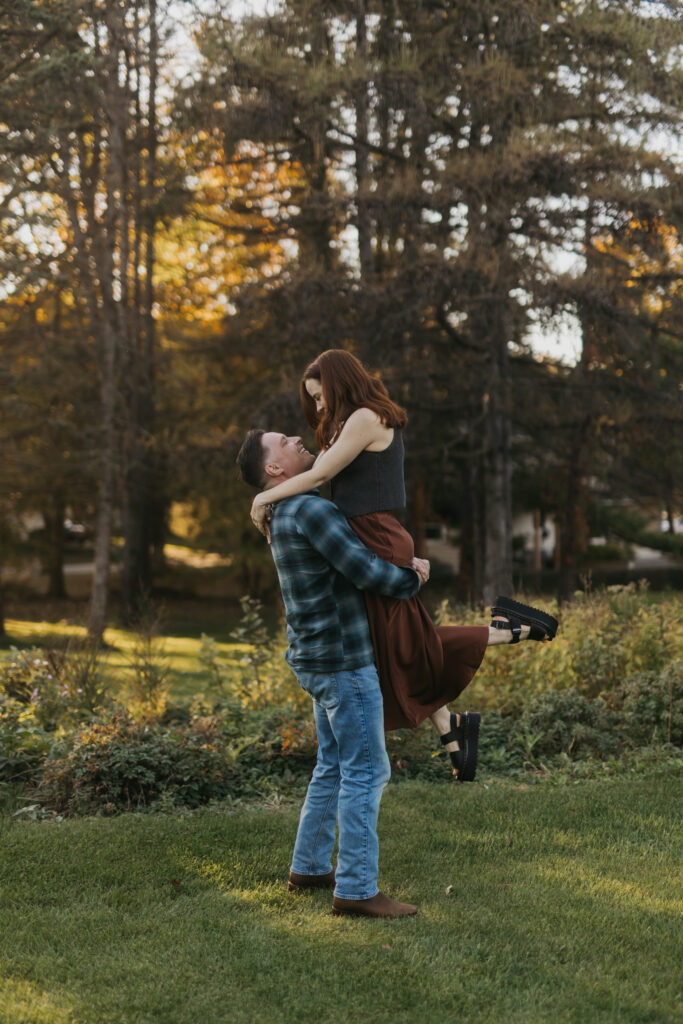 Man lifting his fiancée into a hug, both laughing, on a grassy lawn