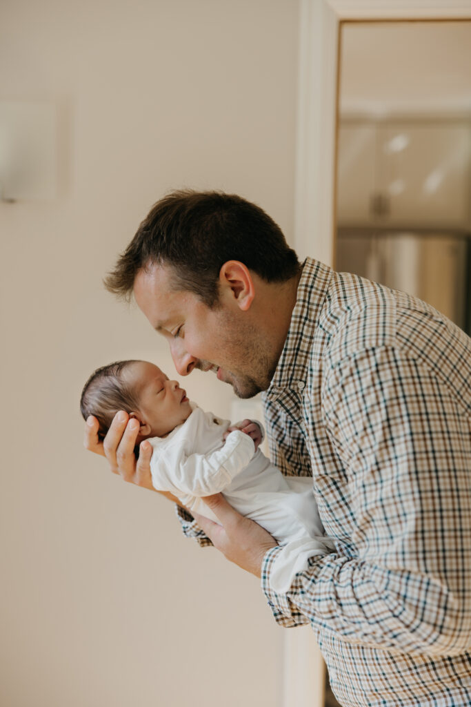 Dad holds his newborn in both hands, face to face, smiling during an at-home newborn session.