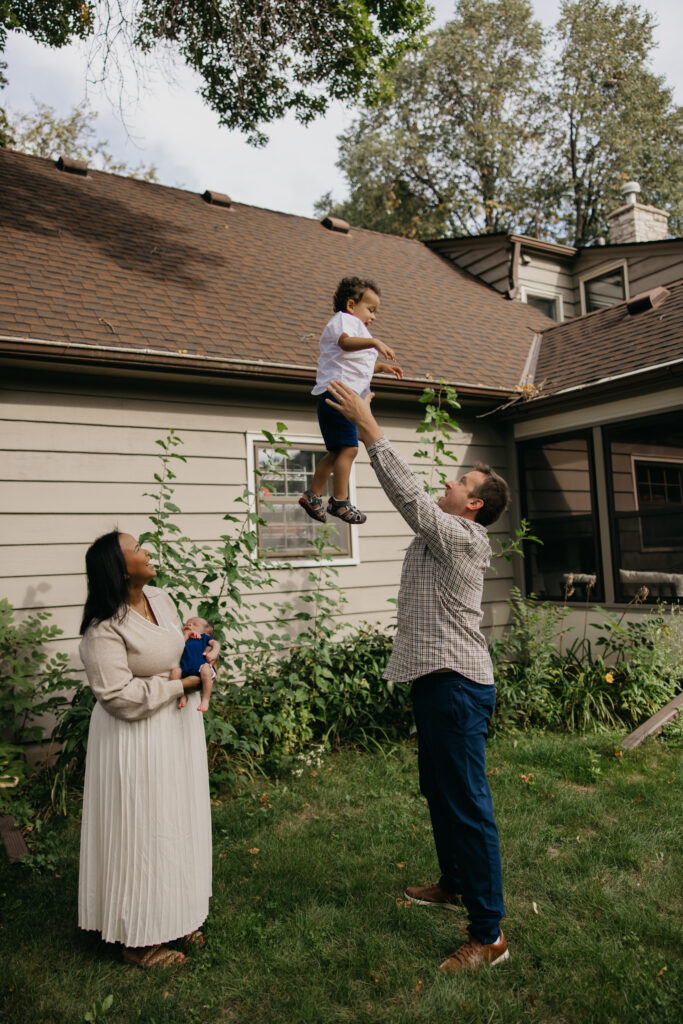 Dad tosses his giggling toddler into the air as mom cradles the newborn during backyard family photos.