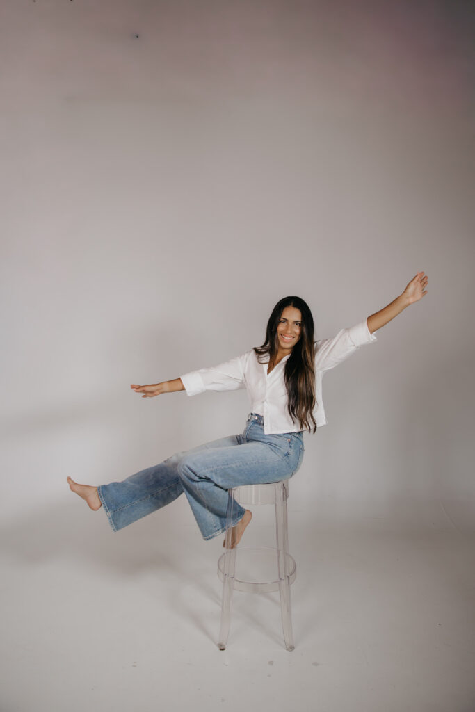 Tatiana Budney in a white button-down and jeans, playfully balancing on a stool in studio